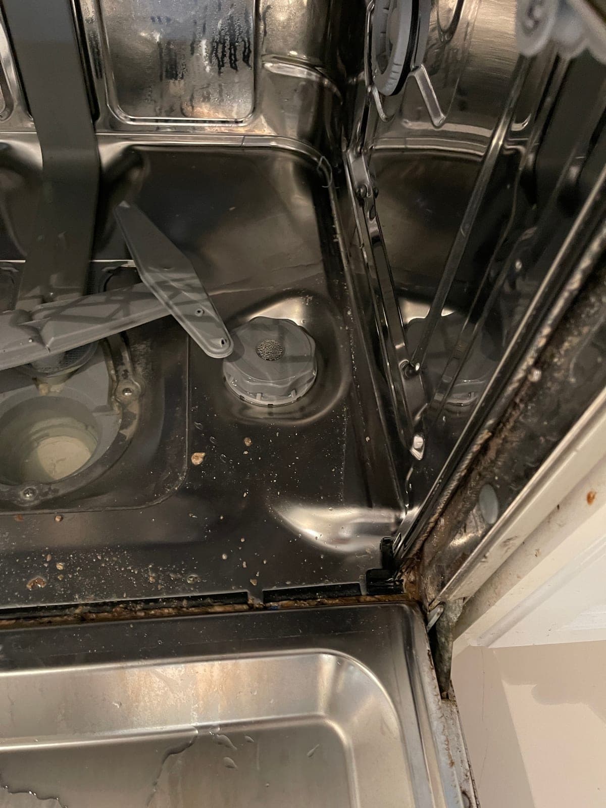 The base of a dishwasher tub before cleaning, with debris around the filter.